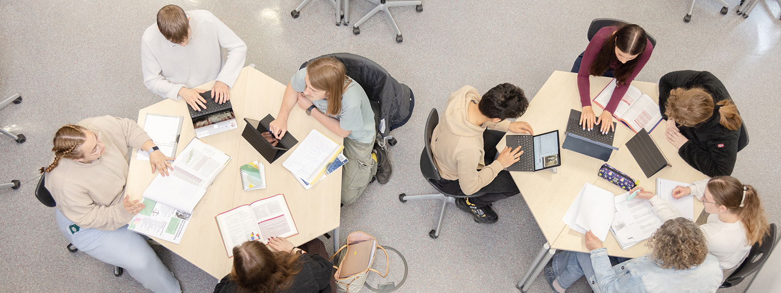 Schüler/-innen arbeiten an verschiedenen Tischen mit Heften, Büchern und Laptops